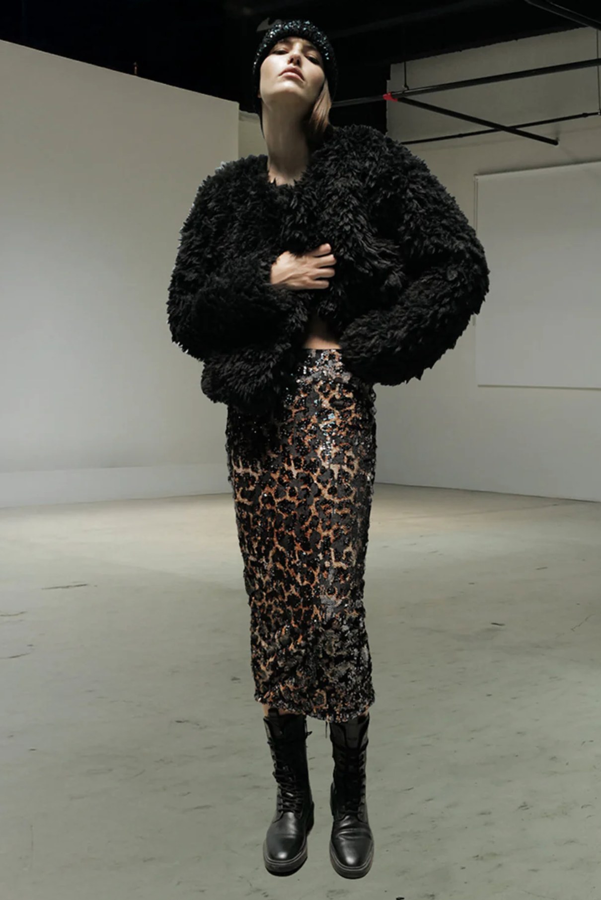 A person wearing the Le Superbe Liza Skirt, a black textured faux fur jacket, black lace-up boots, and a black headband stands in a minimalist industrial space.