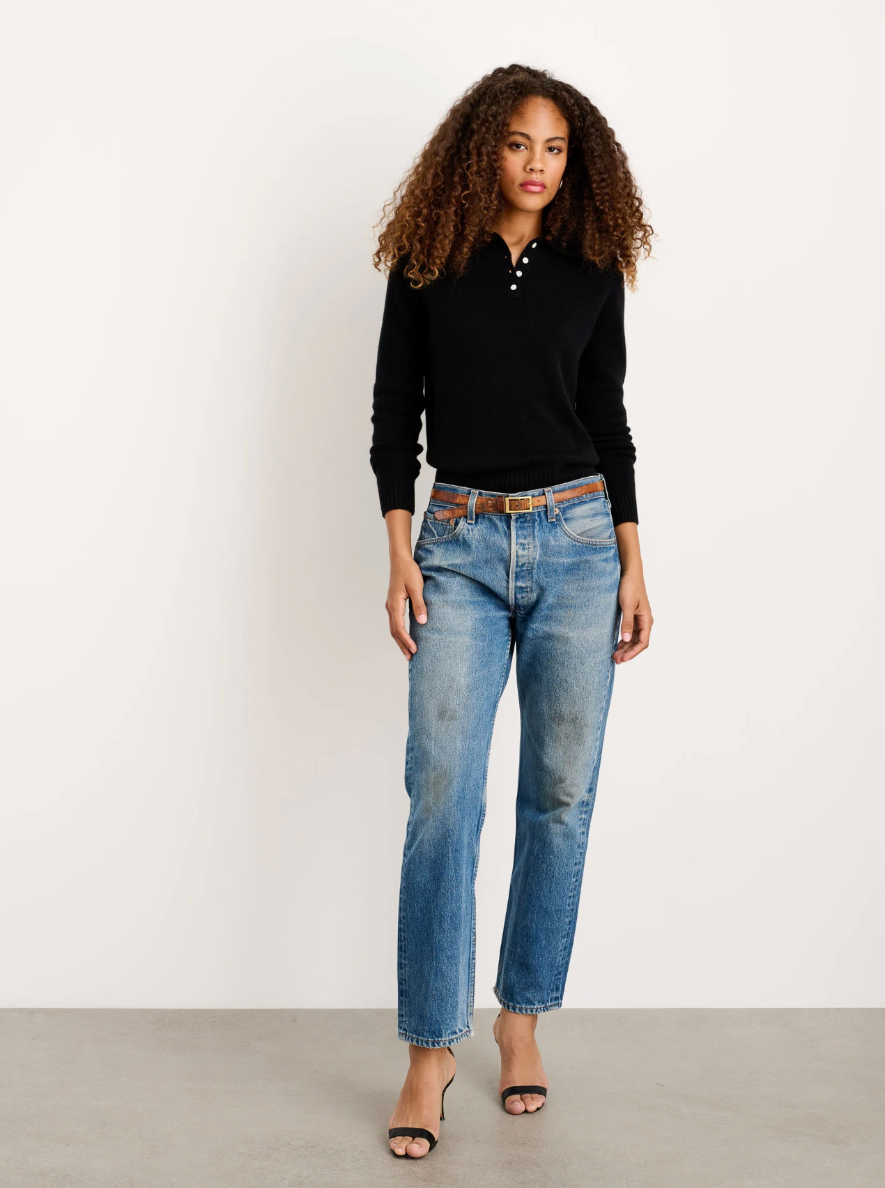 A woman with curly hair wears the Alex Mill Alice Polo Cashmere sweater, blue jeans with a brown belt, and black open-toe heels. She stands on a gray floor against a white wall, looking confidently at the camera.