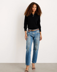 A woman with curly hair wears the Alex Mill Alice Polo Cashmere sweater, blue jeans with a brown belt, and black open-toe heels. She stands on a gray floor against a white wall, looking confidently at the camera.