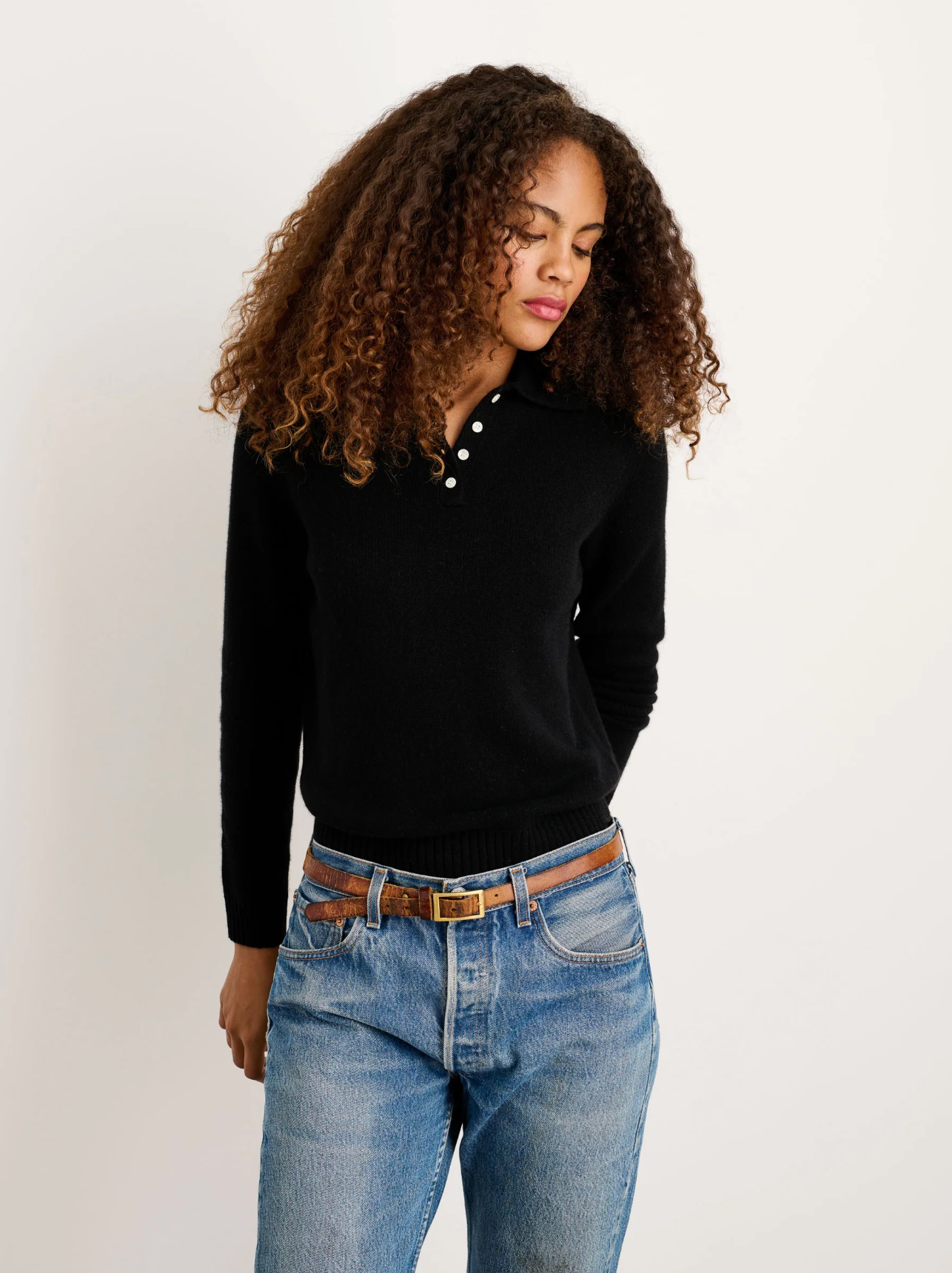 A woman with curly hair wears the Alex Mill Alice Polo Cashmere sweater in black and blue jeans, standing against a white background with her hands behind her back, looking down.