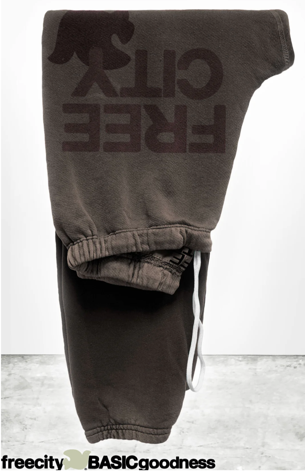 A pair of FREECITY Large Sweatpant by Free City (sparrow, LLC), unisex and dark brown, made in the USA with "FREE CITY" and a bird logo printed boldly, is folded on concrete. "freecity BASICgoodness" appears at the bottom.
