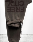 A pair of FREECITY Large Sweatpant by Free City (sparrow, LLC), unisex and dark brown, made in the USA with "FREE CITY" and a bird logo printed boldly, is folded on concrete. "freecity BASICgoodness" appears at the bottom.