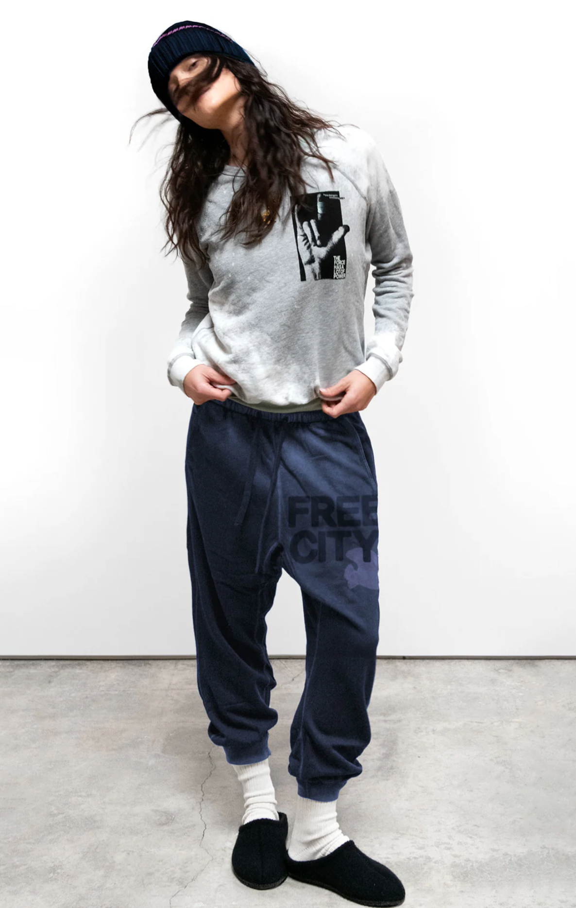 Wearing a beanie, grey graphic sweatshirt, blue FREECITY LARGE SUNFADES pocket sweatpant by Free City (sparrow, LLC), white socks, and black slippers, a person stands against a plain background with head tilted and hands in pockets.