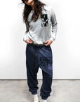 Wearing a beanie, grey graphic sweatshirt, blue FREECITY LARGE SUNFADES pocket sweatpant by Free City (sparrow, LLC), white socks, and black slippers, a person stands against a plain background with head tilted and hands in pockets.