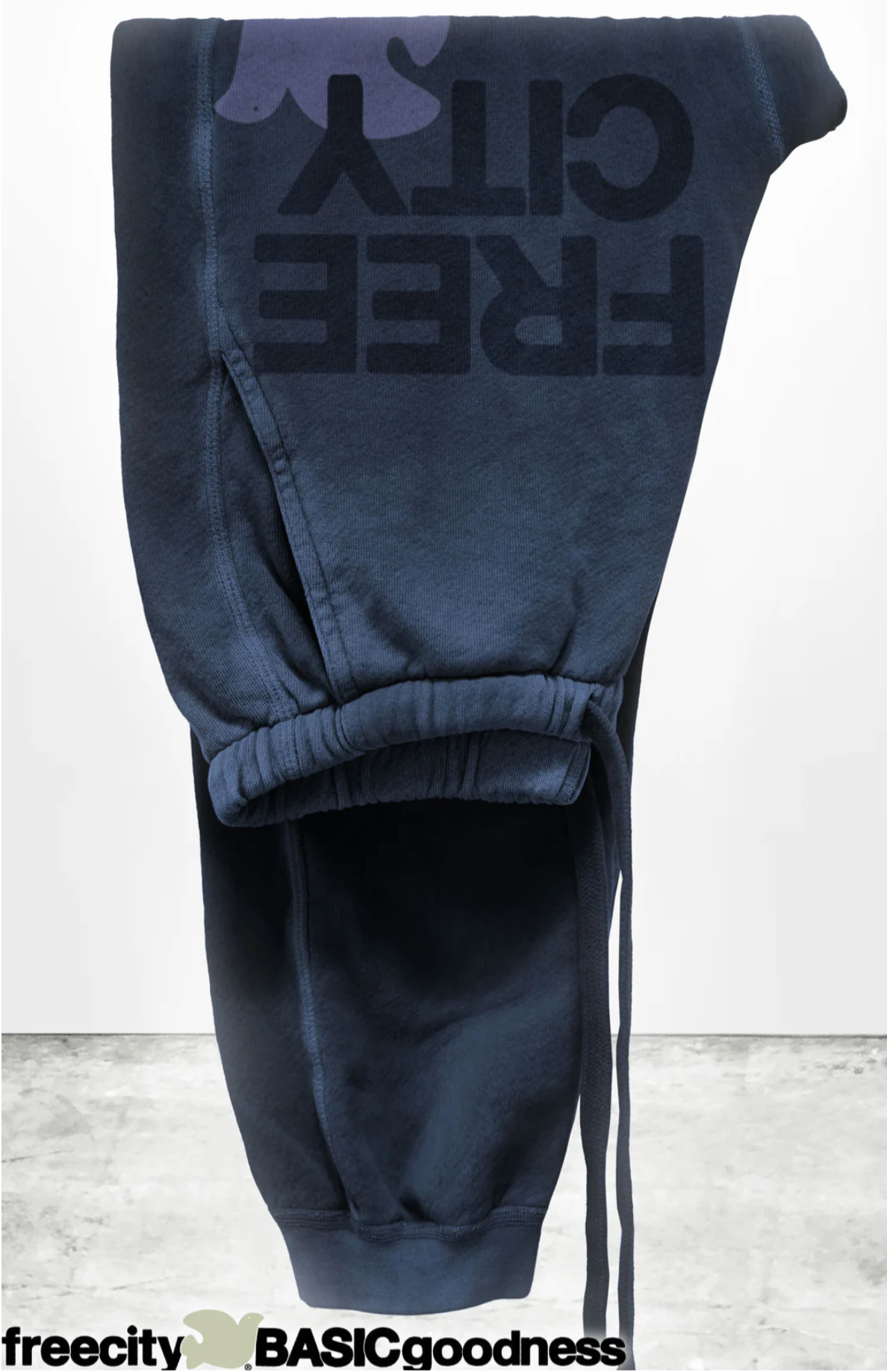 A pair of FREECITY LARGE SUNFADES pocket sweatpants by Free City (sparrow, LLC) is shown upside down in dark blue with "FREE CITY" in bold black letters and a small dove graphic. The background is concrete textured with “freecity BASICgoodness” below.