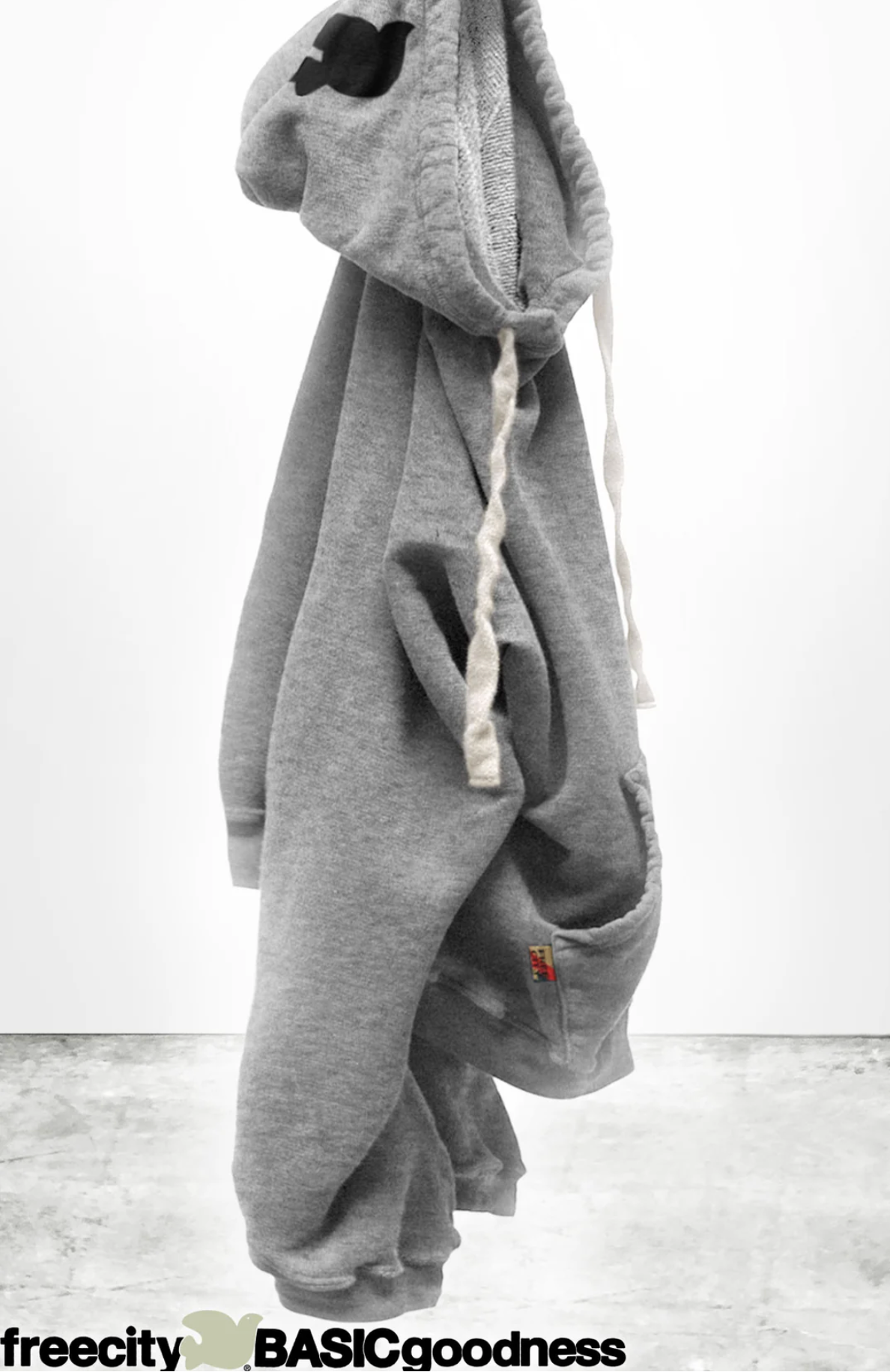 A gray BASIC GOODNESS HOODIE by Free City (sparrow, LLC)—a unisex light French terry pullover with white drawstrings—hangs on a hook against a white background and concrete floor. "freecity BASICgoodness" appears at the bottom.