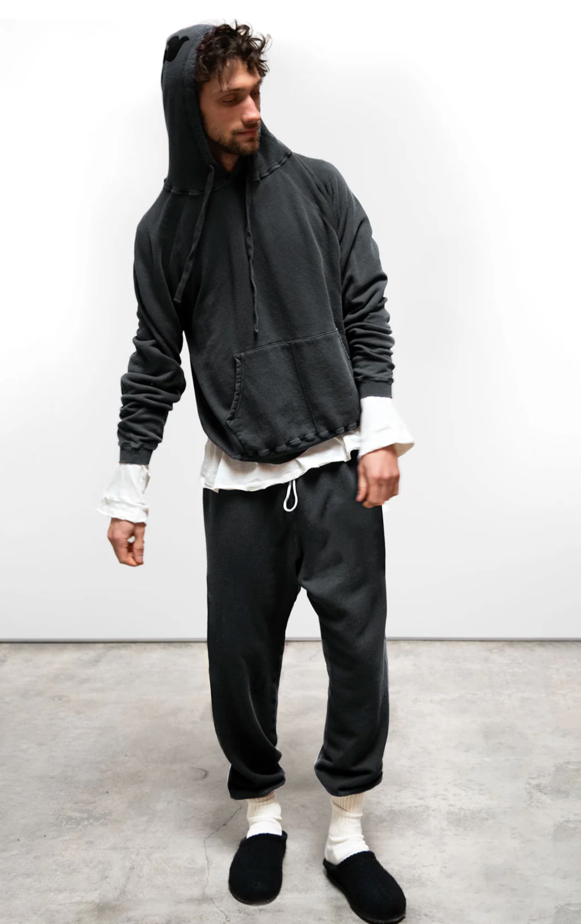 A man wears a loose-fit, made-in-USA Free City BASIC GOODNESS HOODIE with the hood up, matching dark sweatpants, white shirt sleeves, white socks, and black slippers, standing on concrete against a plain white background.