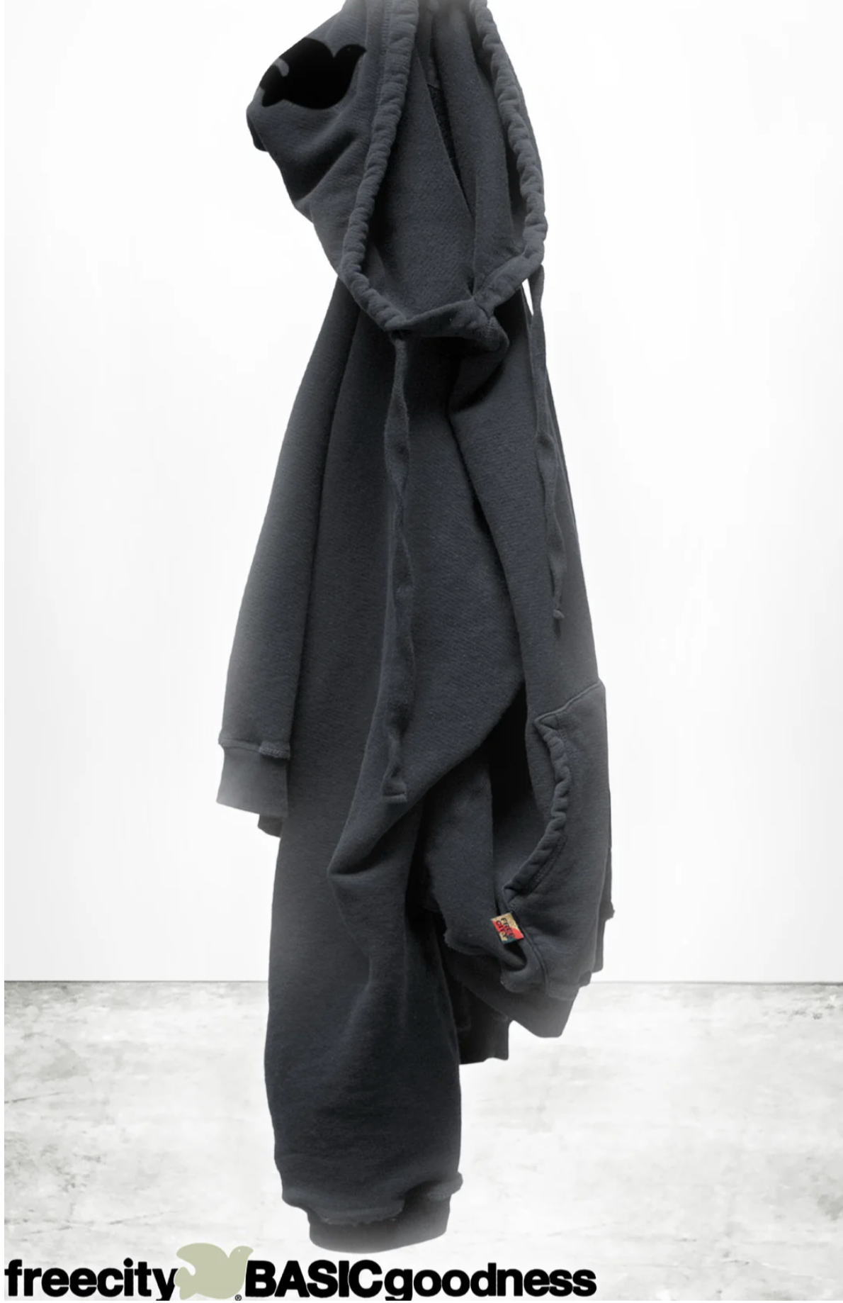 A dark gray unisex BASIC GOODNESS HOODIE by Free City (sparrow, LLC) in light French terry hangs on a hook against a plain white background with a concrete floor. “freecity BASICgoodness” is at the bottom, partially overlapping the image.