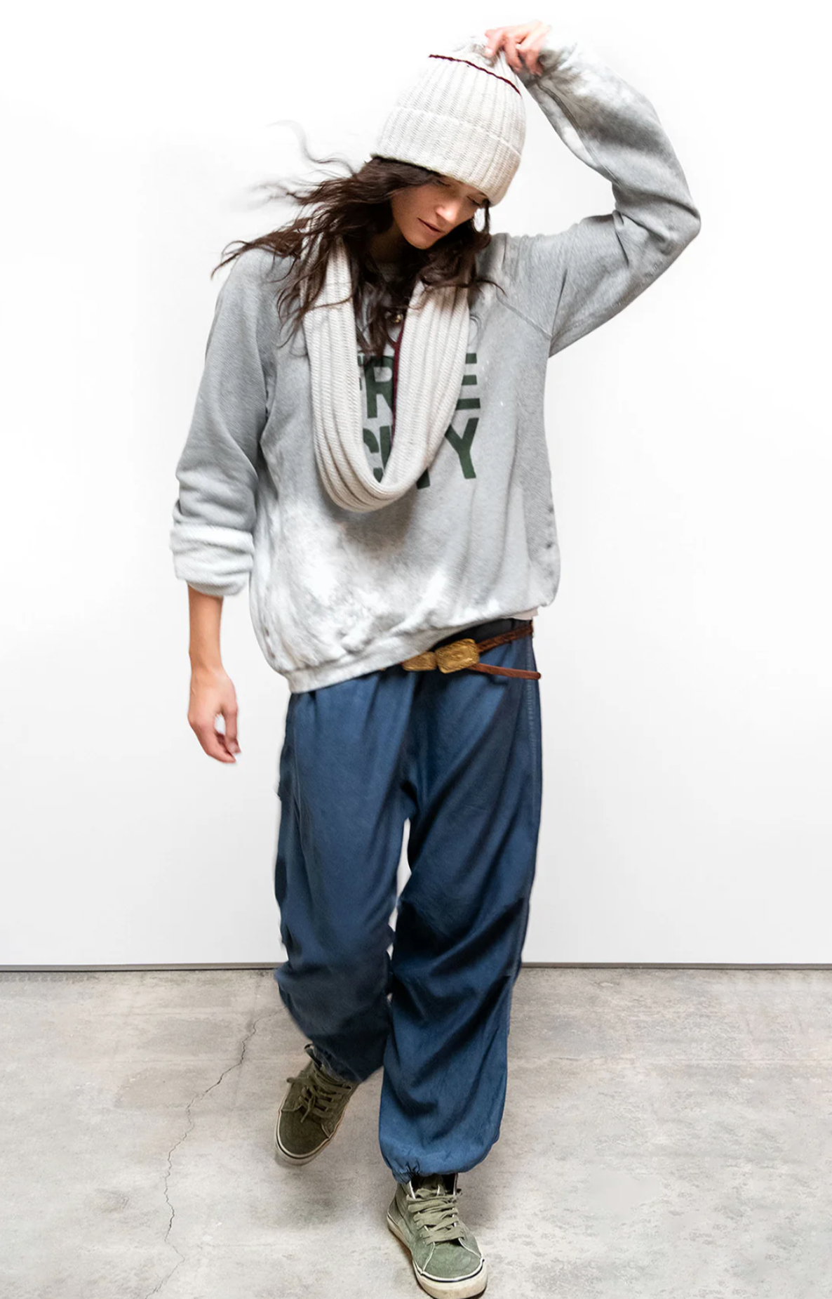 Wearing a gray FREECITY BLEACHOUT RAGLAN by Free City (sparrow, LLC), wide blue pants, green sneakers, and a beige scarf, a person walks on concrete with head down, adjusting their hat.