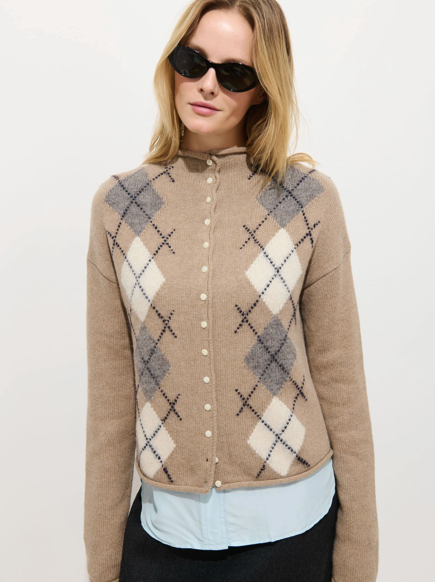 A woman in black sunglasses wears the Alex Mill Taylor Cardigan Argyle with pearl-like buttons over a light blue roll-neck sweater, standing against a plain background for a relaxed fit look.