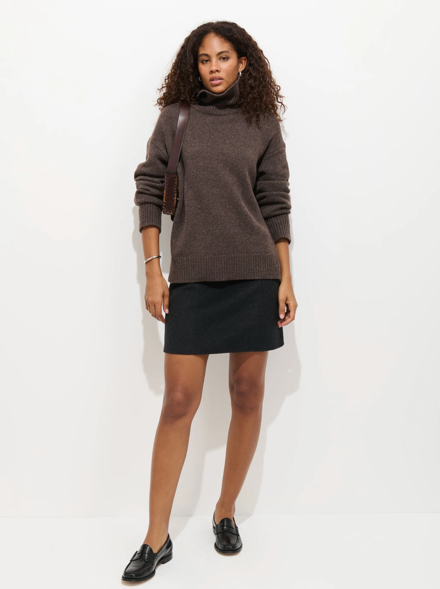 A woman with curly hair wears a dark brown turtleneck, the Alex Mill Naomi Mini Skirt in black with a side zip, and black loafers. She carries a brown shoulder bag and stands against a plain white background.