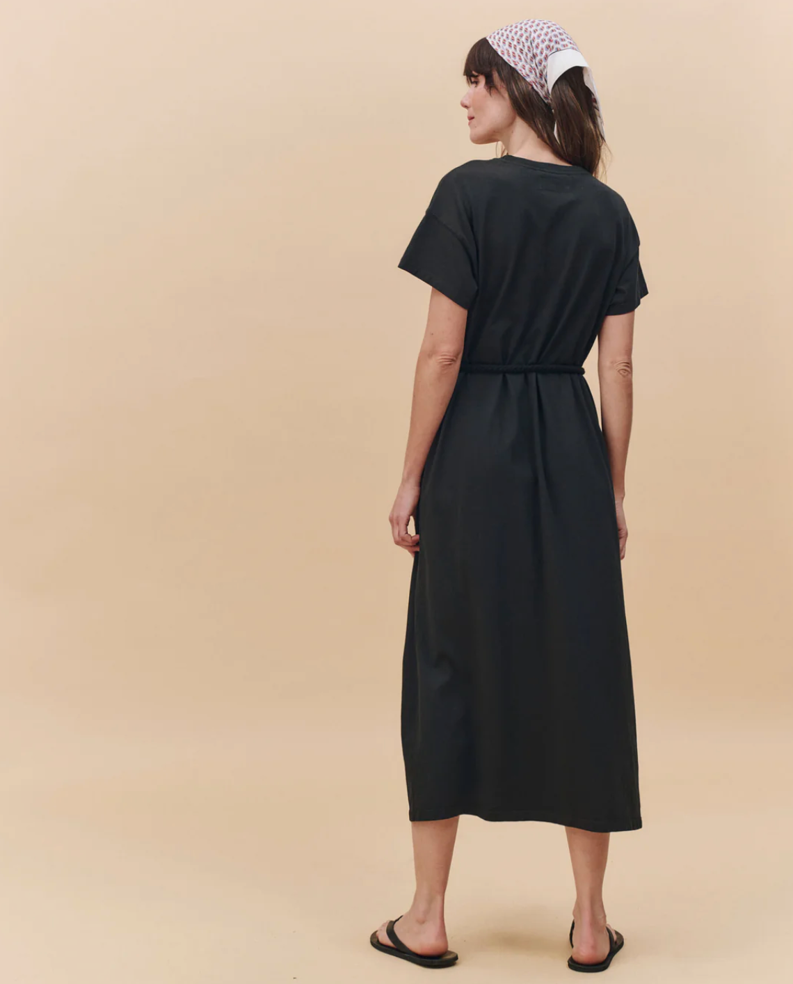 A woman faces away, wearing The Boxy Tee Dress by The Great Inc.—a black crewneck, short-sleeve, ankle-length belted dress—with black sandals and a white mesh headscarf, set against a beige background.