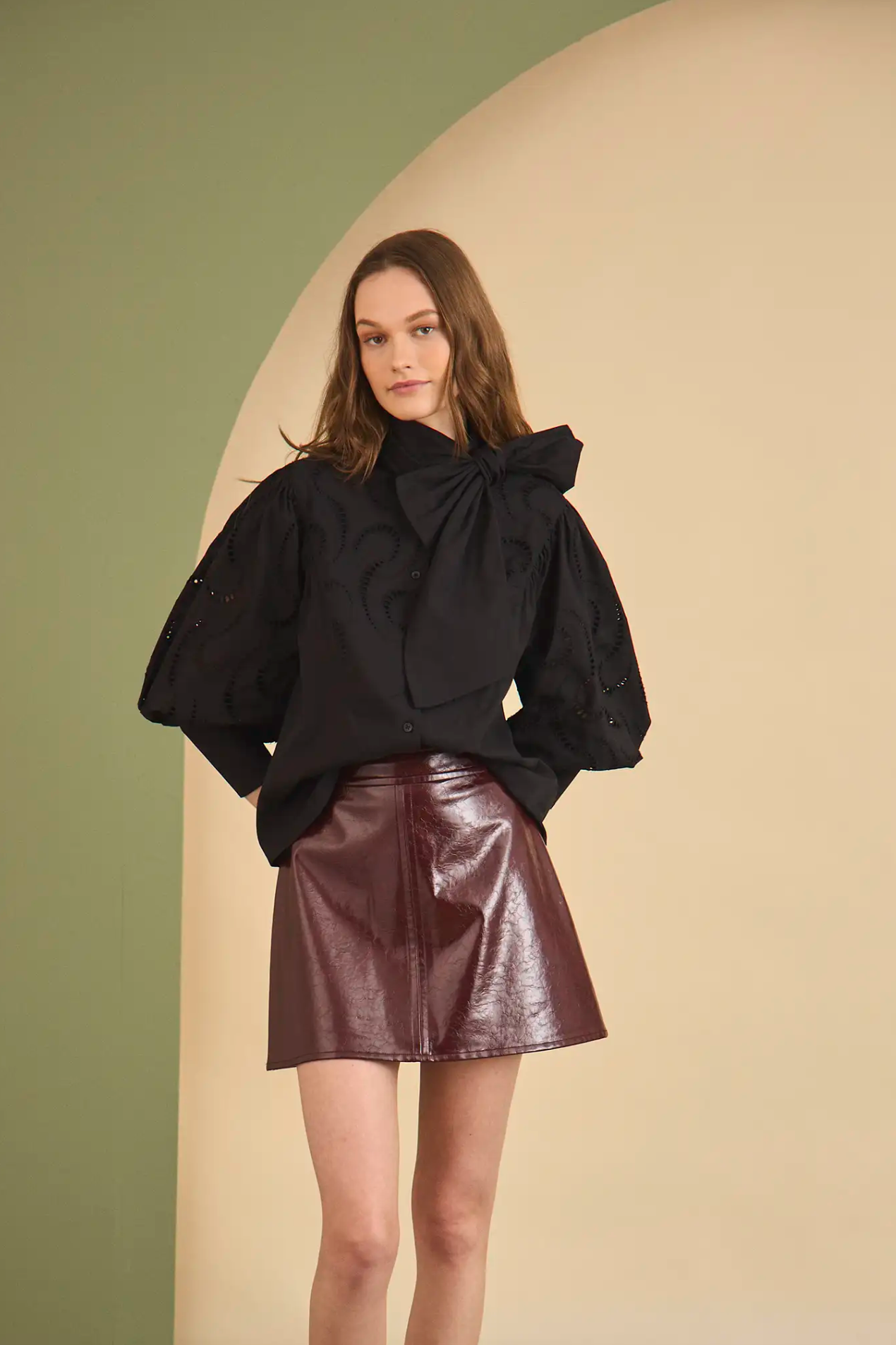 A woman poses against a beige and green backdrop, wearing the Hunter Bell NYC Shay Blouse—featuring spacious sleeves and a dramatic bow collar—paired with a shiny burgundy mini skirt.