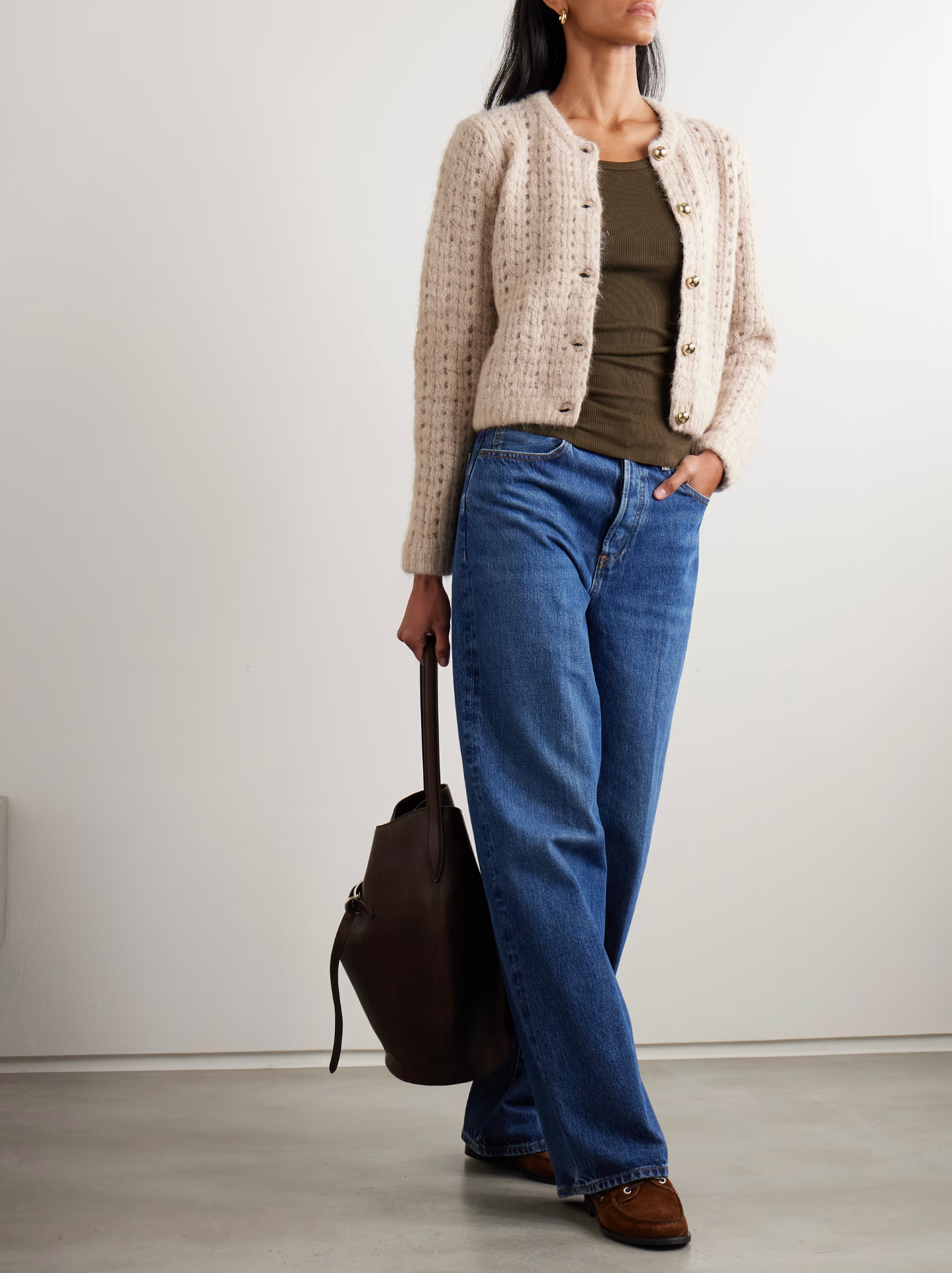 A woman wears FRAME's The Plush Cardi in beige over an olive top with blue wide-leg jeans, brown shoes, and holds a dark brown bag. She stands against a plain wall, her face partially out of frame.