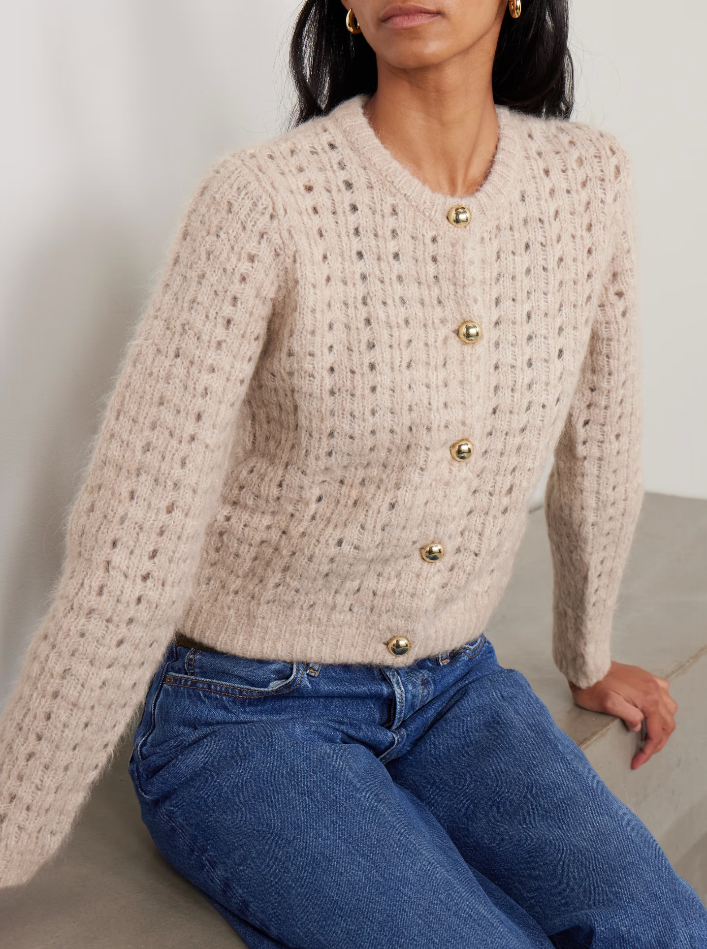 A woman wears FRAME's The Plush Cardi, a soft beige open-weave knit with gold buttons, paired with blue jeans as she sits on a light surface against a neutral background.