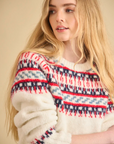 A woman with long blonde hair smiles softly as she models the Josie Sweater by Hunter Bell NYC—a cozy, cream Fairisle knit featuring red, blue, and navy geometric patterns—standing against a neutral background.