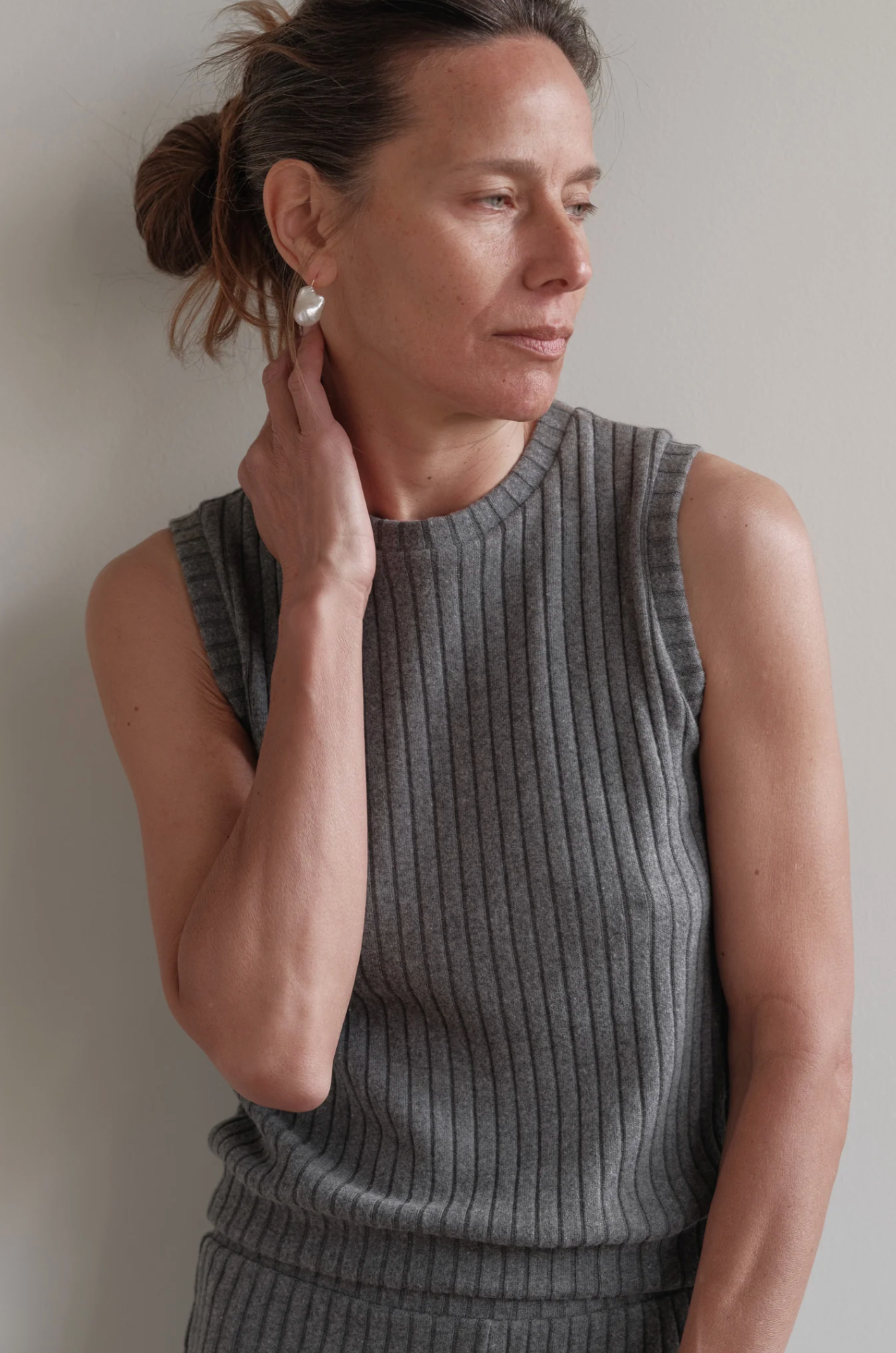 A woman with light skin and brown hair tied back wears The Sweater Rib Shell by Donni, a sleeveless ribbed gray top in a soft Tencel blend, as she touches her ear and looks thoughtfully to the side against a neutral background.