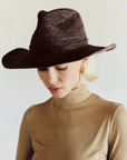 A person with platinum blonde hair wears the Janessa Leone Sacha Chocolate hat, gold hoop earrings, and a beige turtleneck top, looking down against a plain light background.