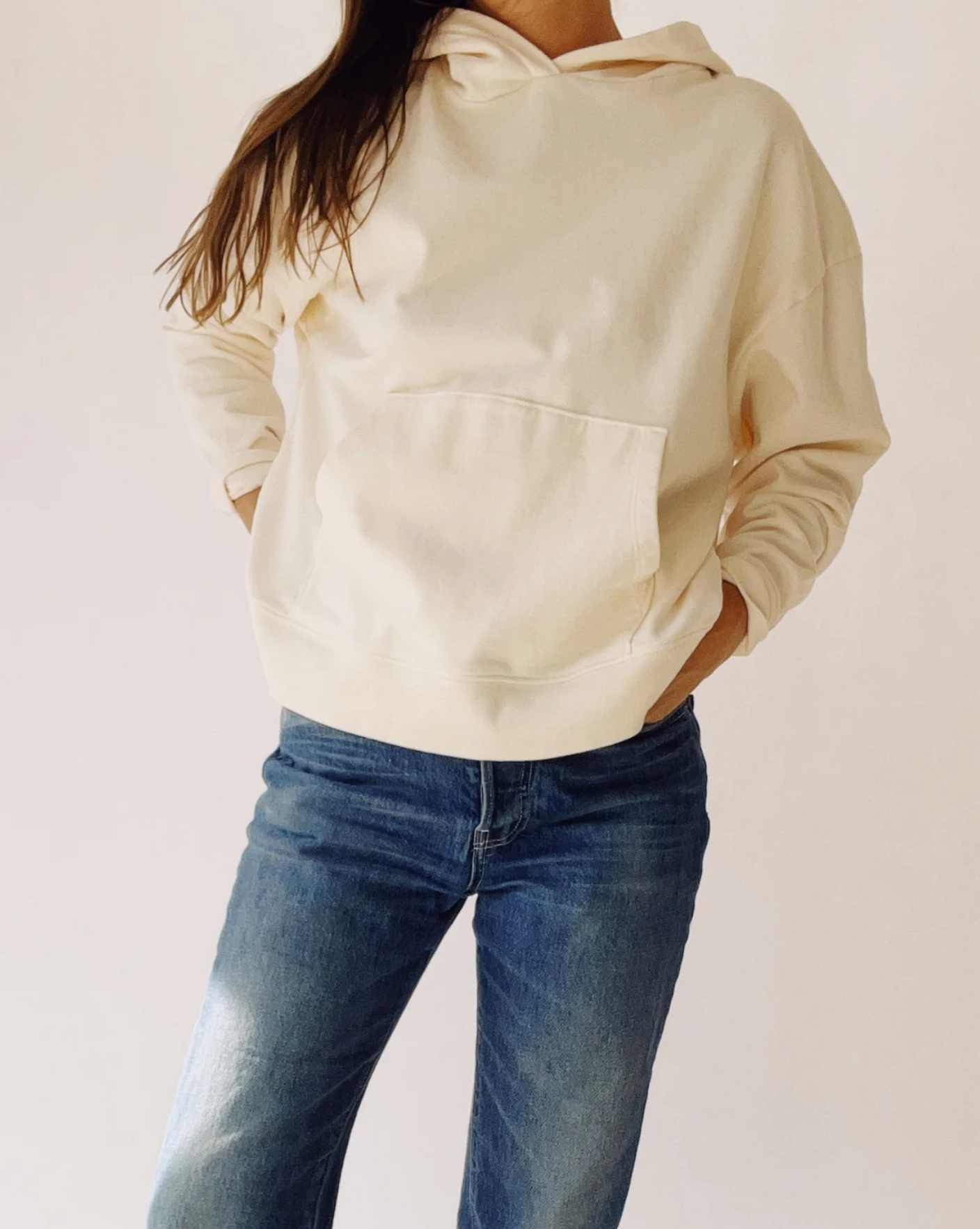 A person models The Hoodie by Hey Gang—a cream-colored unisex French terry cotton hoodie—paired with blue jeans. They stand hands-in-pockets against a plain light background, with their face hidden from view.