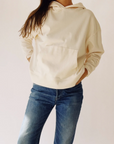 A person models The Hoodie by Hey Gang—a cream-colored unisex French terry cotton hoodie—paired with blue jeans. They stand hands-in-pockets against a plain light background, with their face hidden from view.