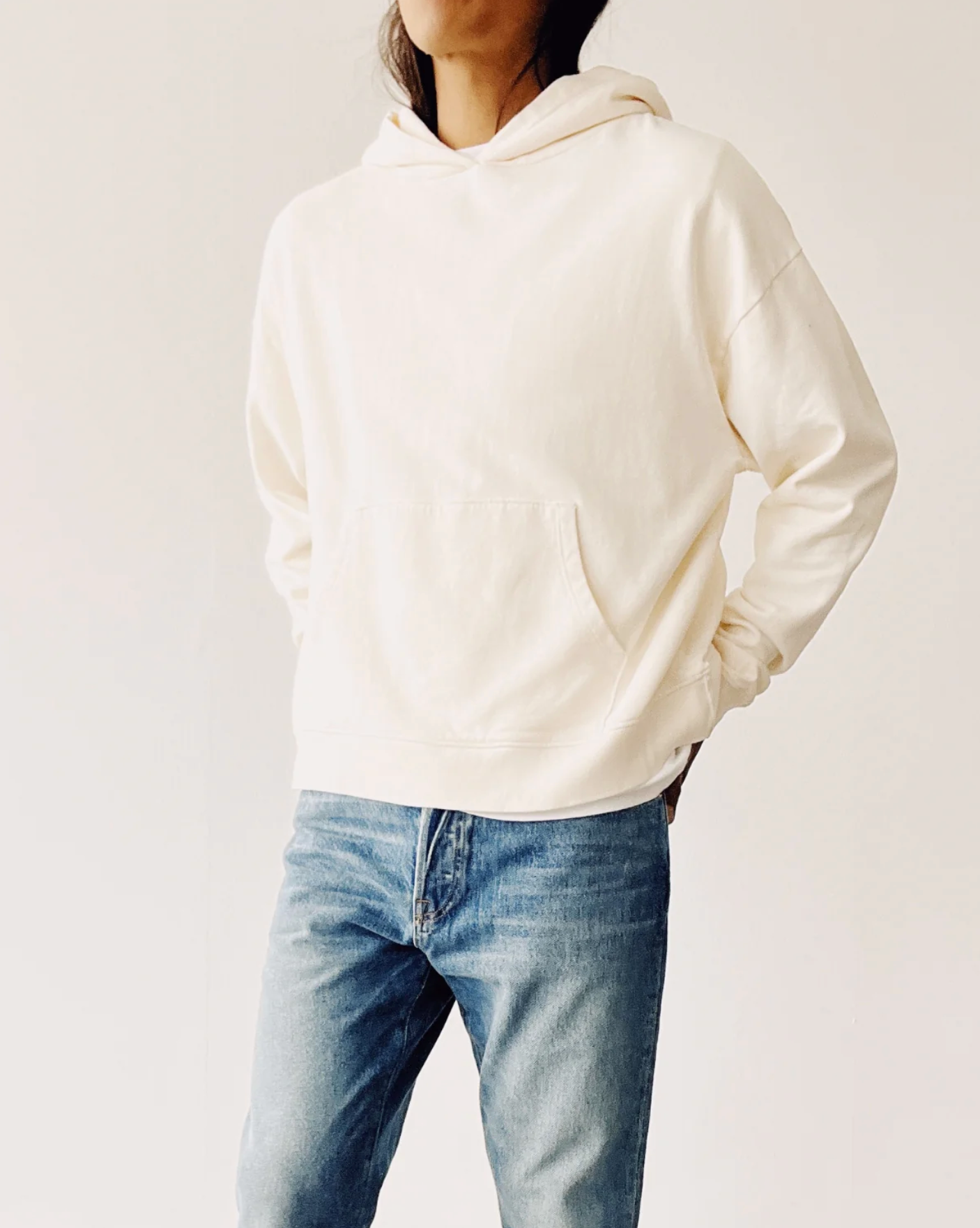 A person wears The Hoodie by Hey Gang—a cream-colored, boxy unisex cotton French terry hoodie with a front pocket—paired with blue jeans, standing against a plain light background, hands in pockets, head partially cropped.