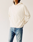 A person wears The Hoodie by Hey Gang—a cream-colored, boxy unisex cotton French terry hoodie with a front pocket—paired with blue jeans, standing against a plain light background, hands in pockets, head partially cropped.