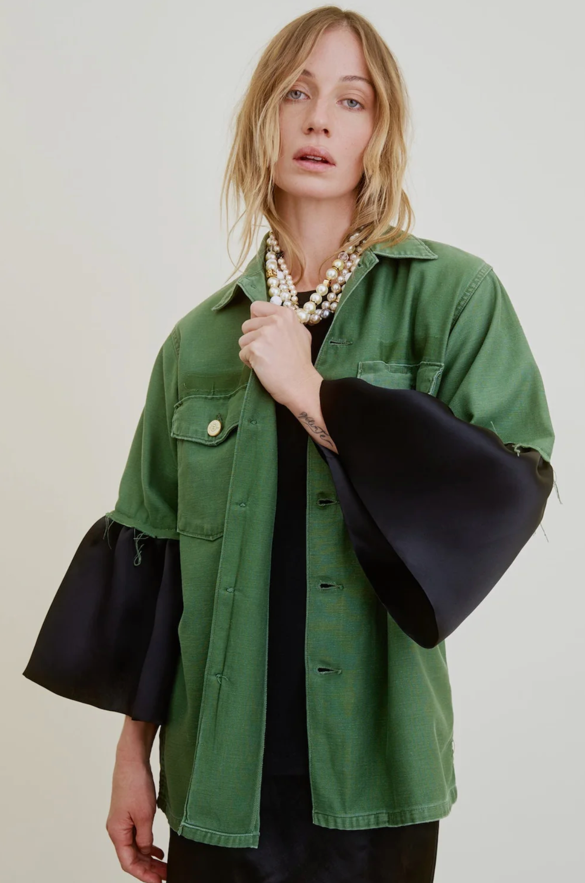 A person with wavy blonde hair models the Aquarius Cocktail Hooper Suiting Cuff Army Jacket—featuring designer buttons and wide black sleeves—over a black top and chunky pearl necklace, set against a plain off-white background.
