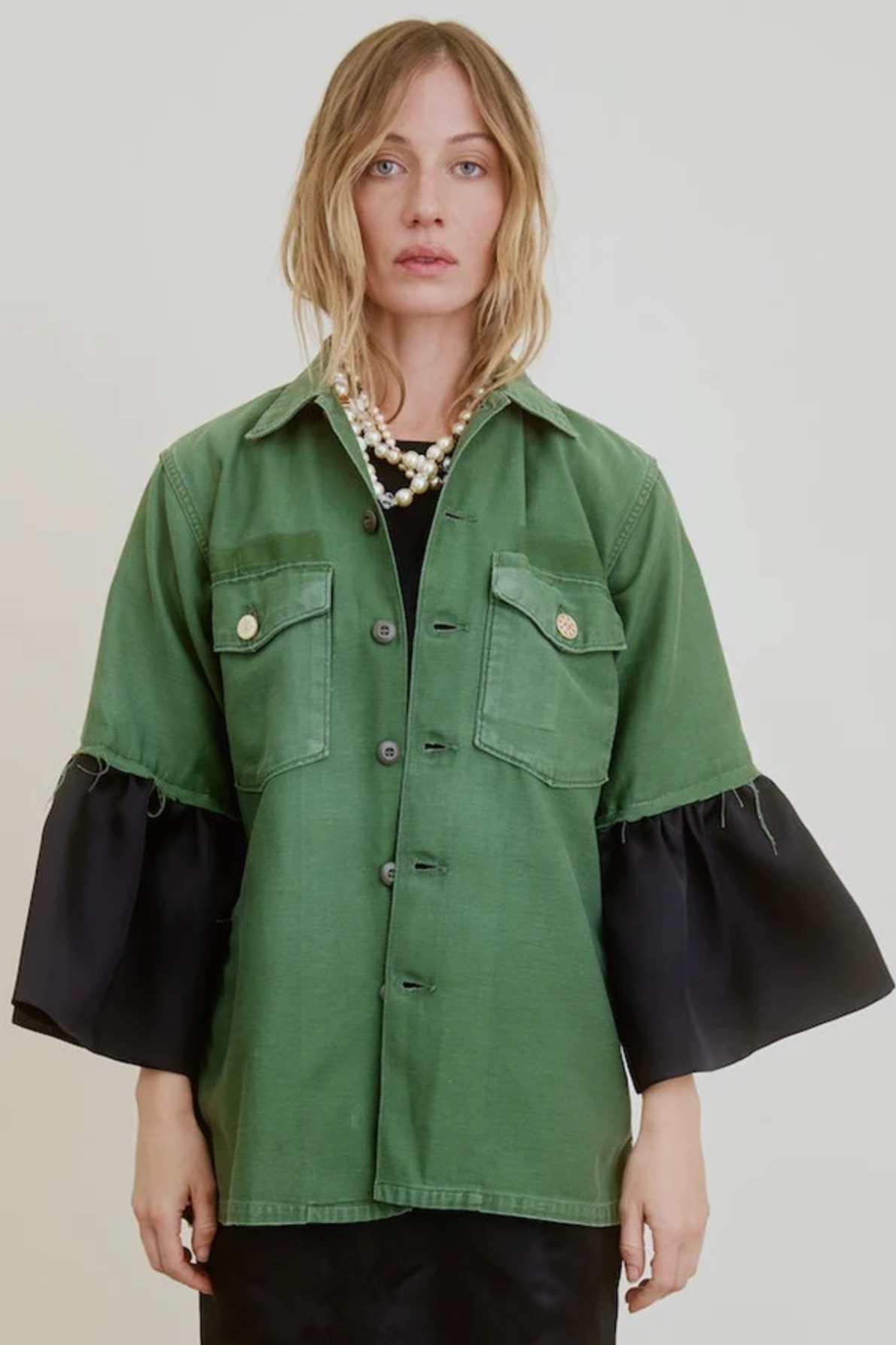 A woman with wavy blonde hair wears the Aquarius Cocktail Hooper Suiting Cuff Army Jacket, styled with a pearl necklace and black undershirt, standing against a plain light background.