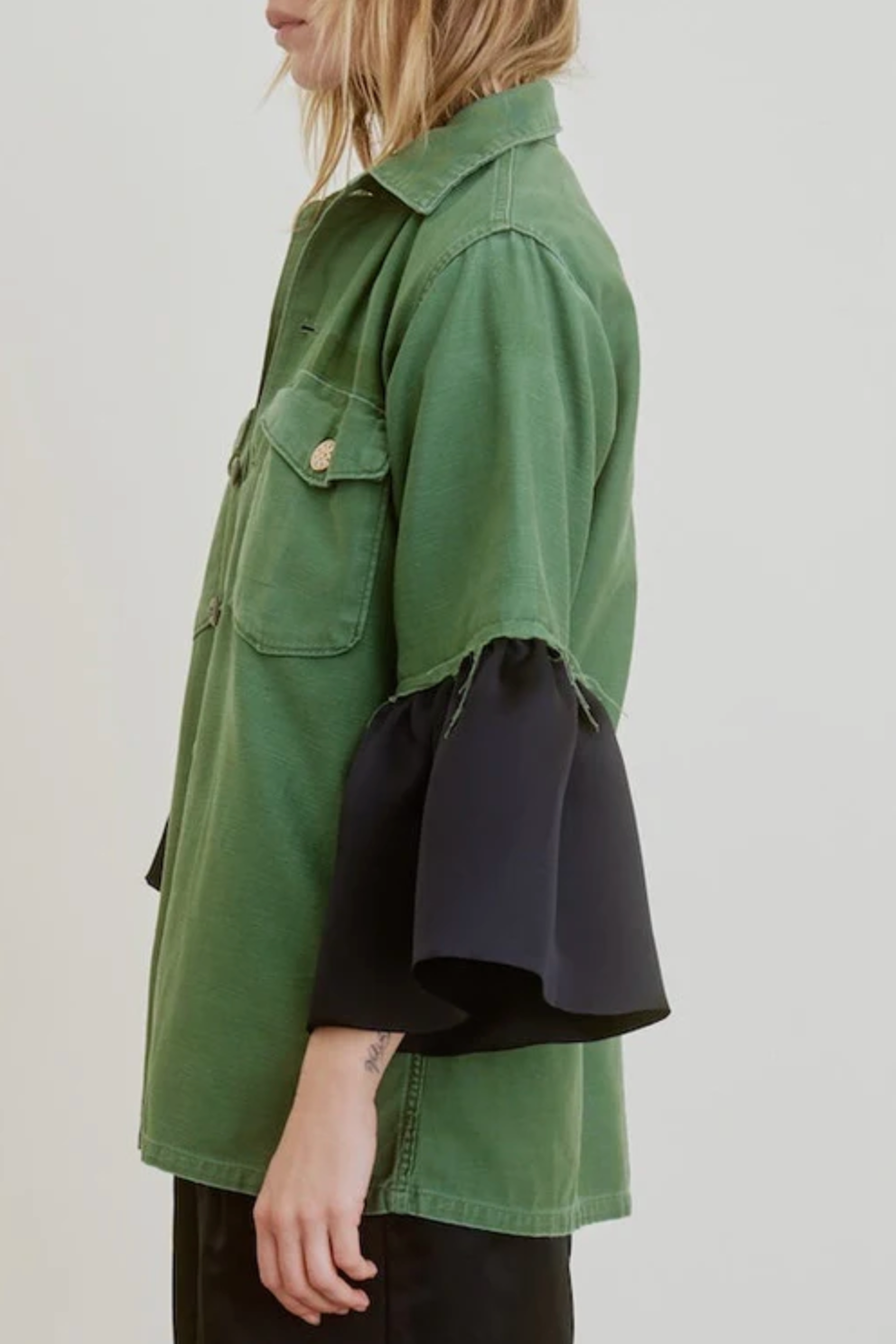 A person wearing the Aquarius Cocktail Hooper Suiting Cuff Army Jacket, a green button-up with chest pockets, designer buttons, and unique black bell-shaped sleeve cuffs, shown from the side against a plain background.