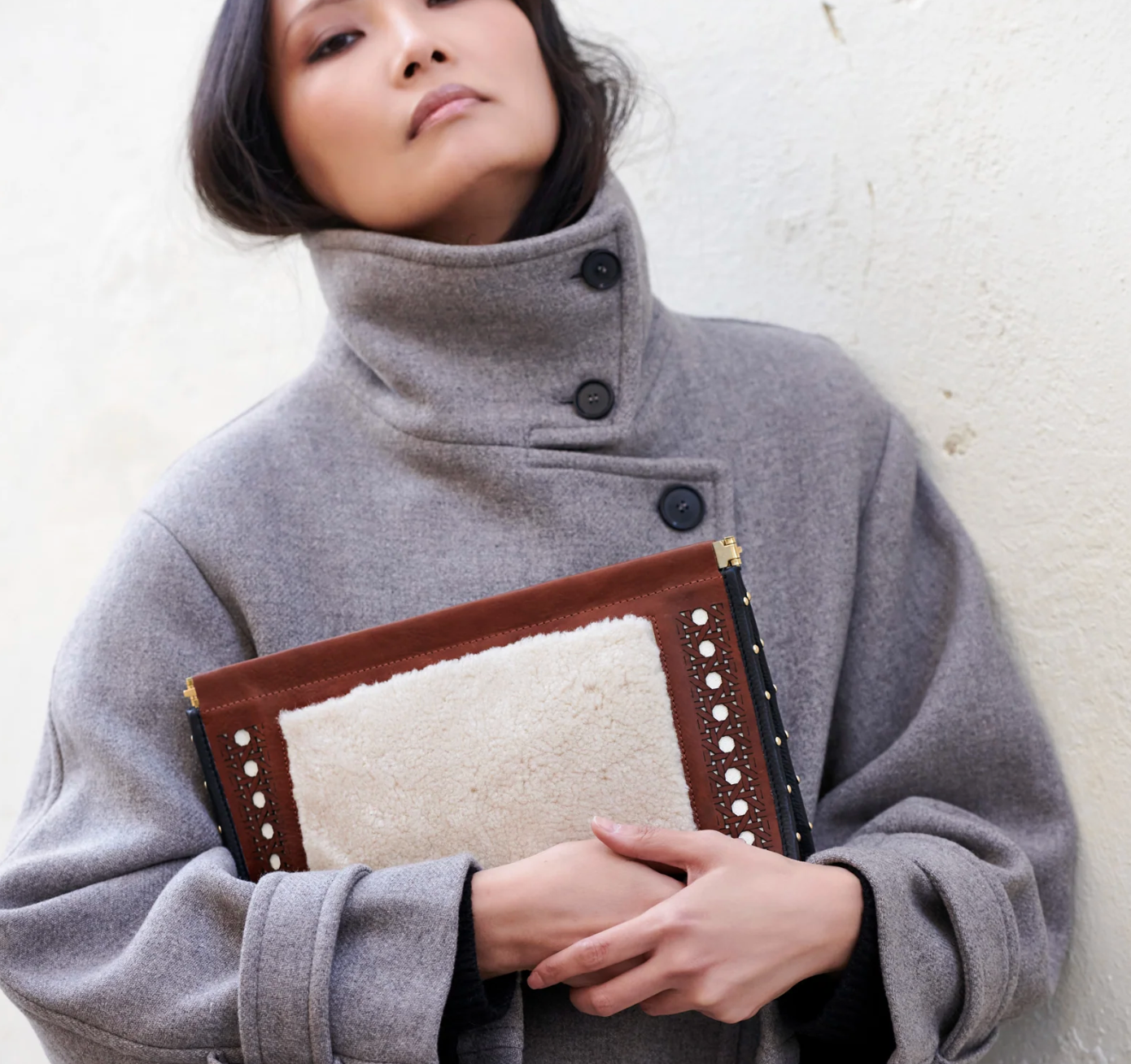 A woman in a high-collared gray coat holds the Kempton & Co. Snap Clutch - Leather Rattan, standing against a light textured wall.