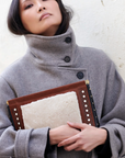 A woman in a high-collared gray coat holds the Kempton & Co. Snap Clutch - Leather Rattan, standing against a light textured wall.