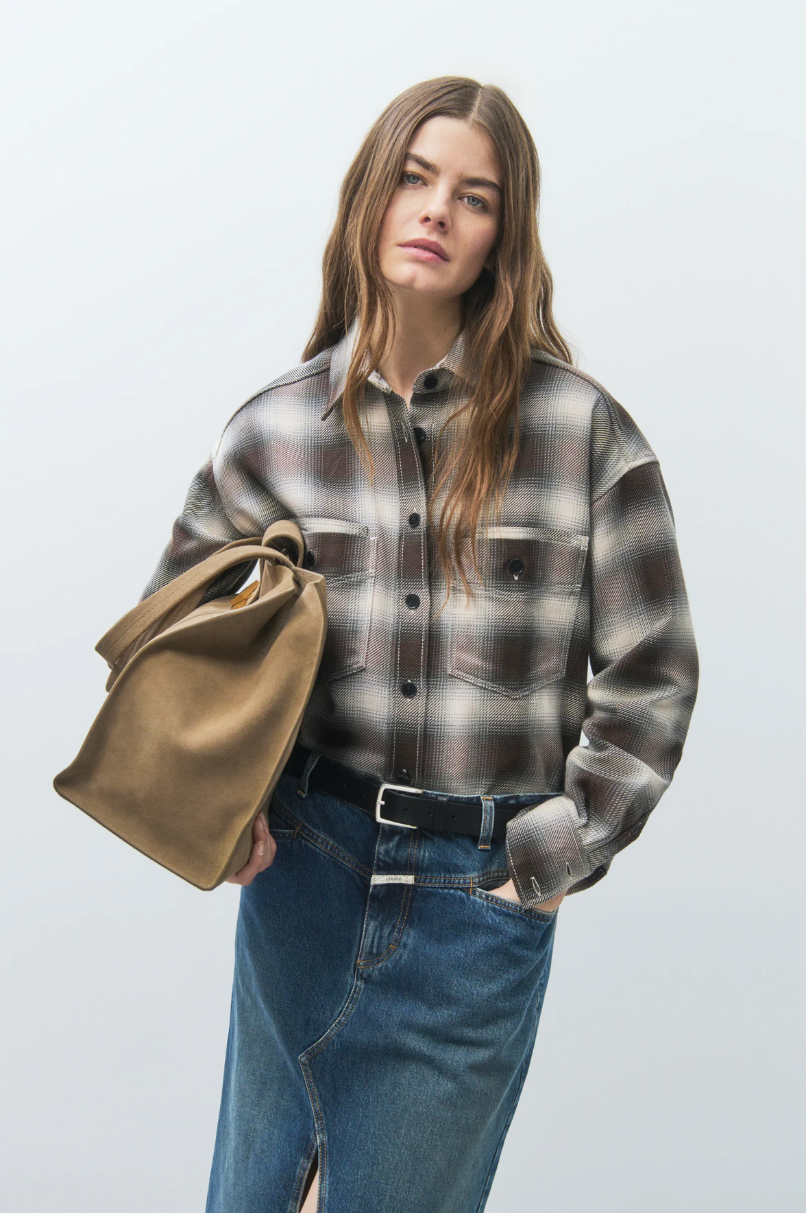 A woman with long, wavy brown hair wears the CLOSED Cropped Front OS Shirt and a denim skirt with a front slit, holding a large tan handbag. She stands against a plain light background, confidently facing the camera.