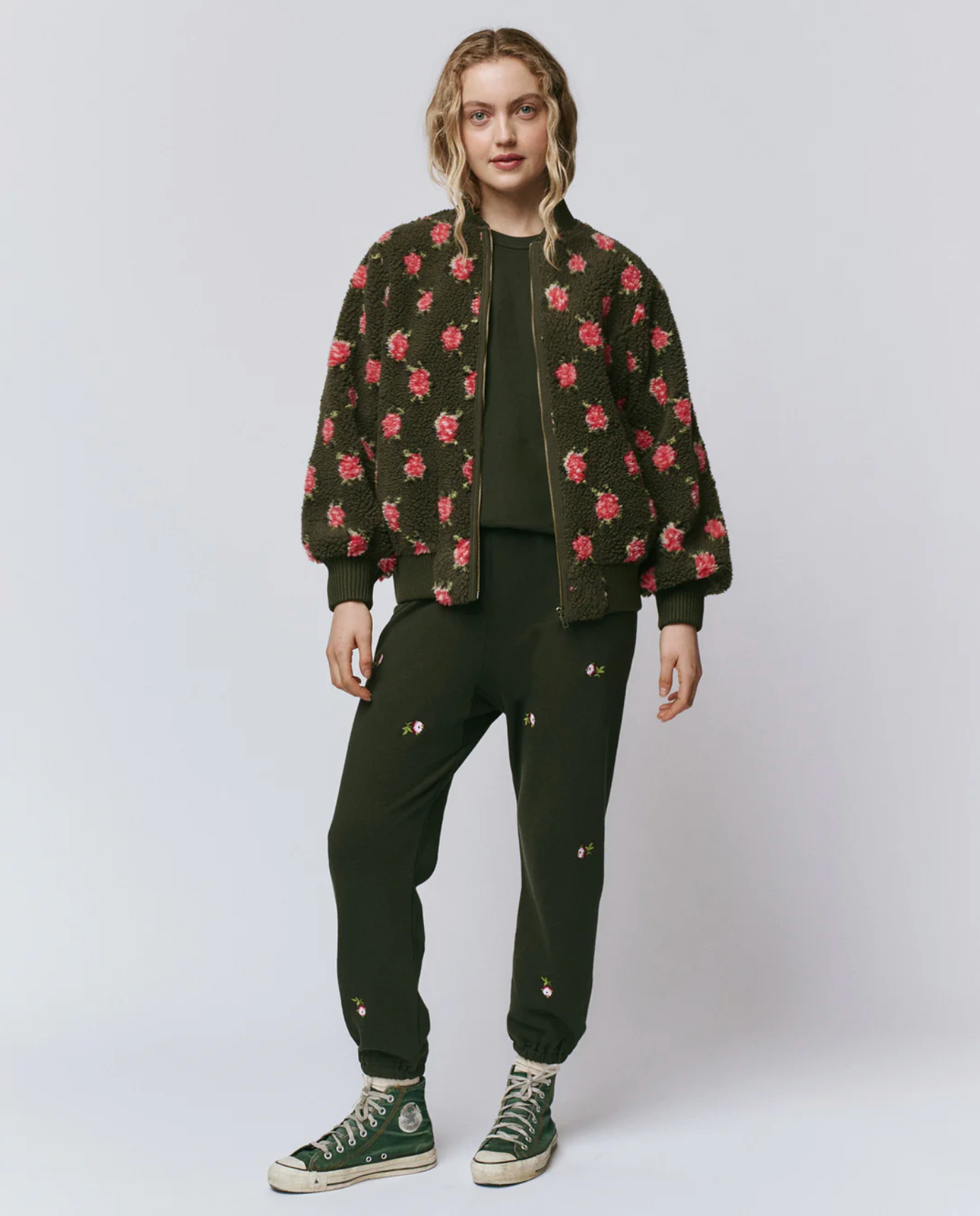 A person stands against a plain background wearing an olive green floral-embroidered outfit, green high-top sneakers, and The Great Inc. Zip Front Plush Bomber with a floral texture. Their wavy hair is worn loose.