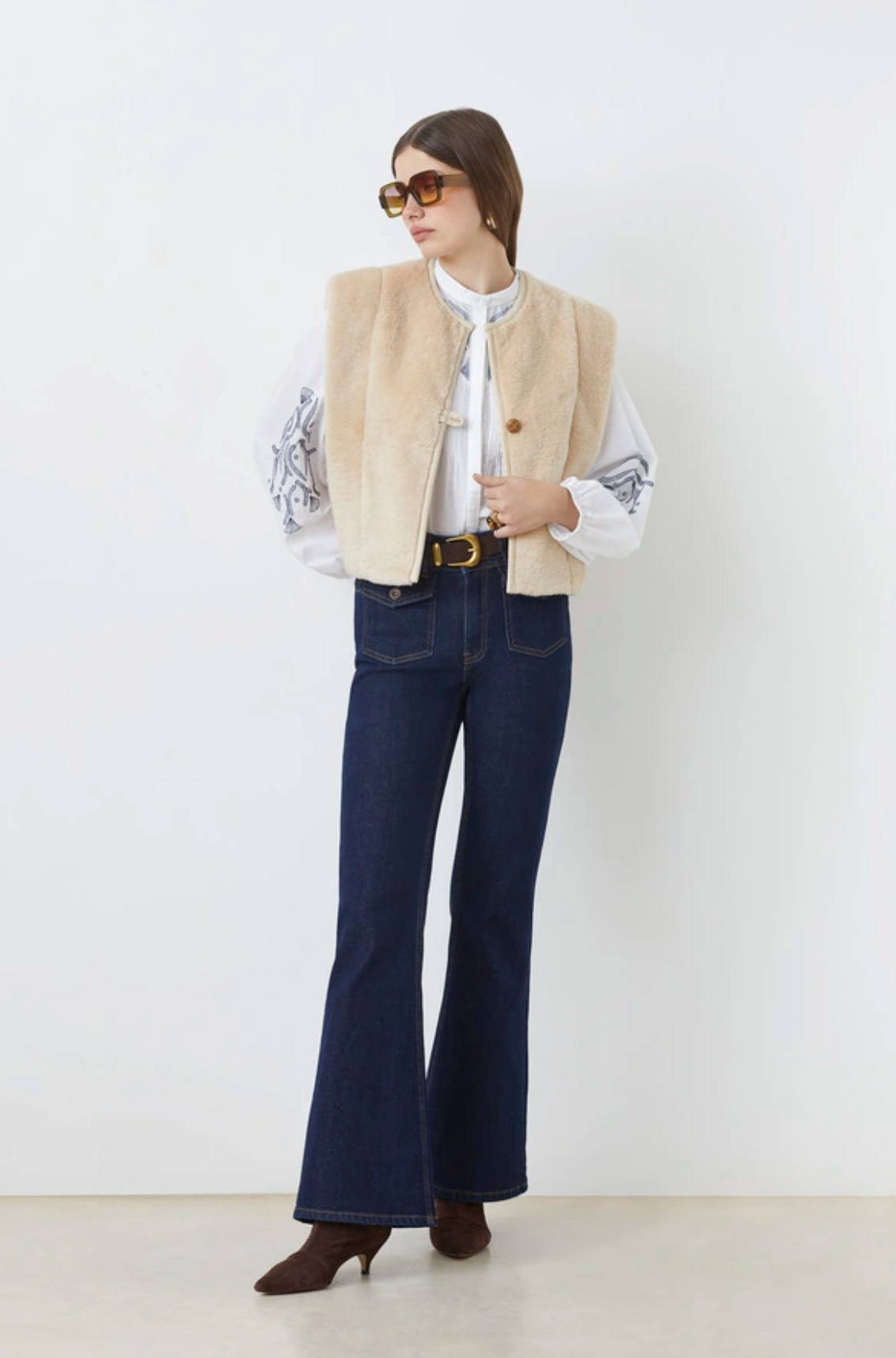 A woman stands against a white background, wearing sunglasses, the SUNCOO Diego Vest—a sleeveless faux fur jacket with beige piping—over a white embroidered blouse, high-waisted dark blue flared jeans, and brown pointed-toe boots.