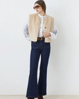 A woman stands against a white background, wearing sunglasses, the SUNCOO Diego Vest—a sleeveless faux fur jacket with beige piping—over a white embroidered blouse, high-waisted dark blue flared jeans, and brown pointed-toe boots.
