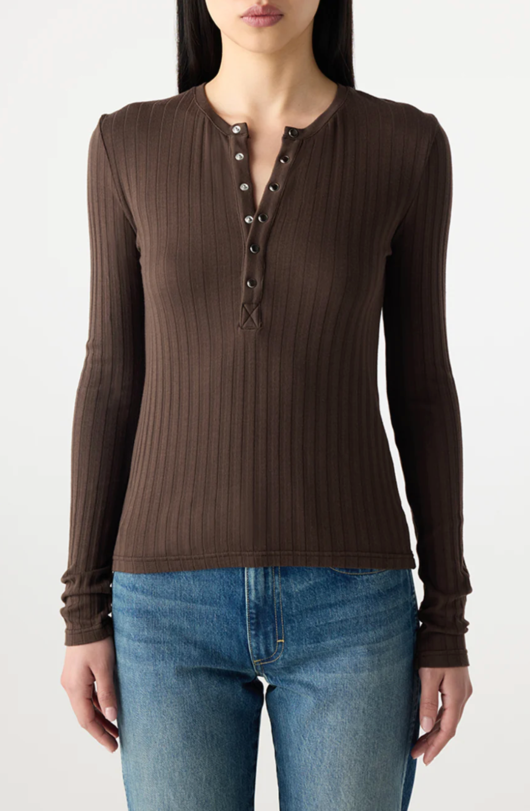 A woman wears the AMO Denim Lottie Top, a fitted ribbed henley in brown with metal buttons, paired with blue jeans against a light background. This cotton blend top is perfect for everyday style.