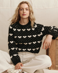 A woman with wavy blonde hair wears The Heart Crew sweater by The Great Inc. with off-white pants, seated cross-legged on the floor, resting one arm on a wooden box against a neutral fabric backdrop.