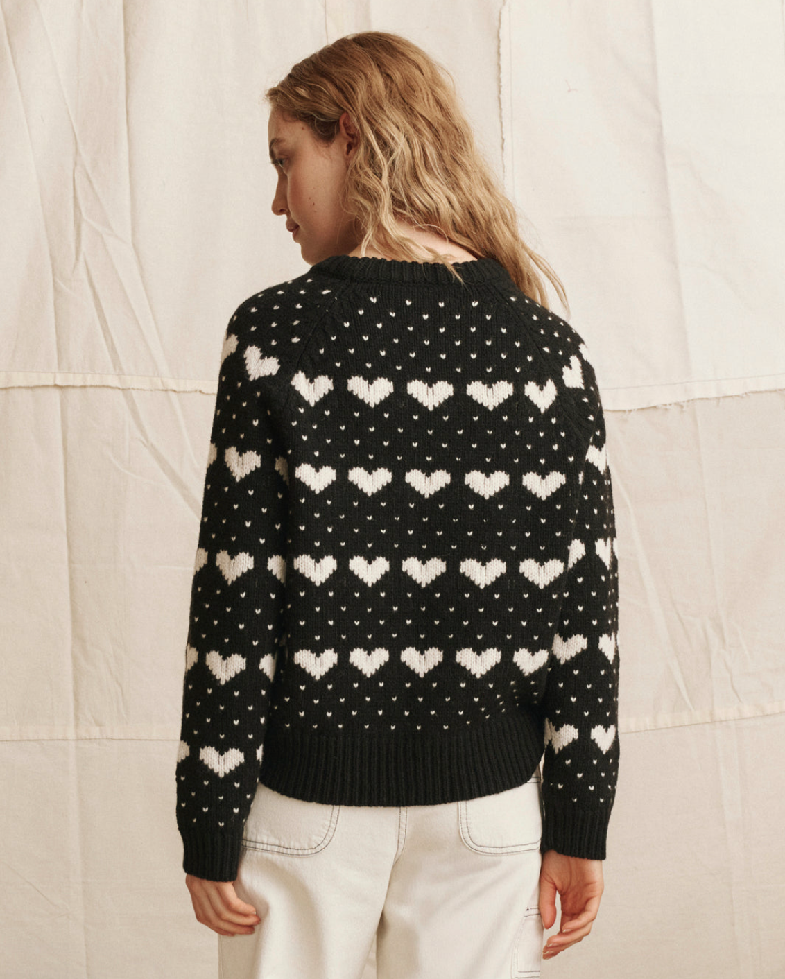 Someone with wavy, light brown hair models The Great Inc.'s The Heart Crew—a vintage-style black sweater with white hearts and dots—paired with light pants, against a neutral textured fabric backdrop.