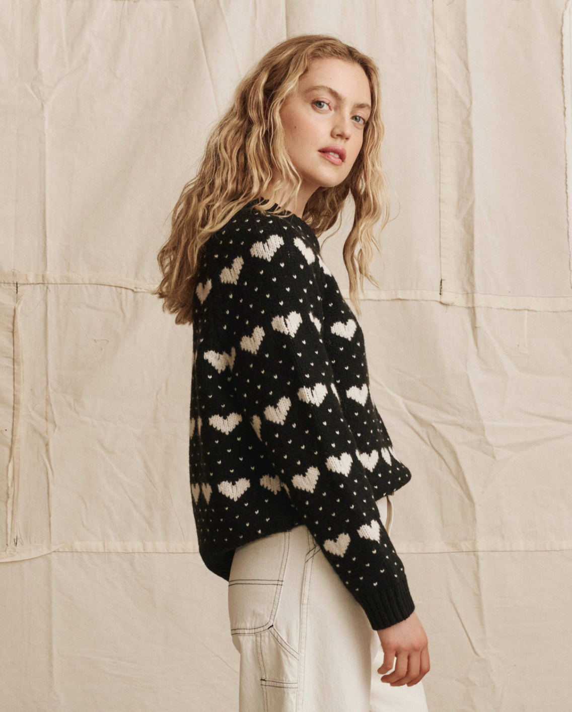 A young woman with wavy blonde hair wears The Heart Crew sweater by The Great Inc. and white pants, standing sideways and looking at the camera against a beige, textured fabric background.