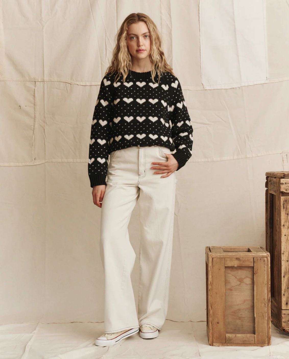 A woman stands before a cream-colored fabric backdrop wearing The Heart Crew sweater by The Great Inc., paired with wide-leg white pants and white shoes. Wooden crates are stacked to her right.