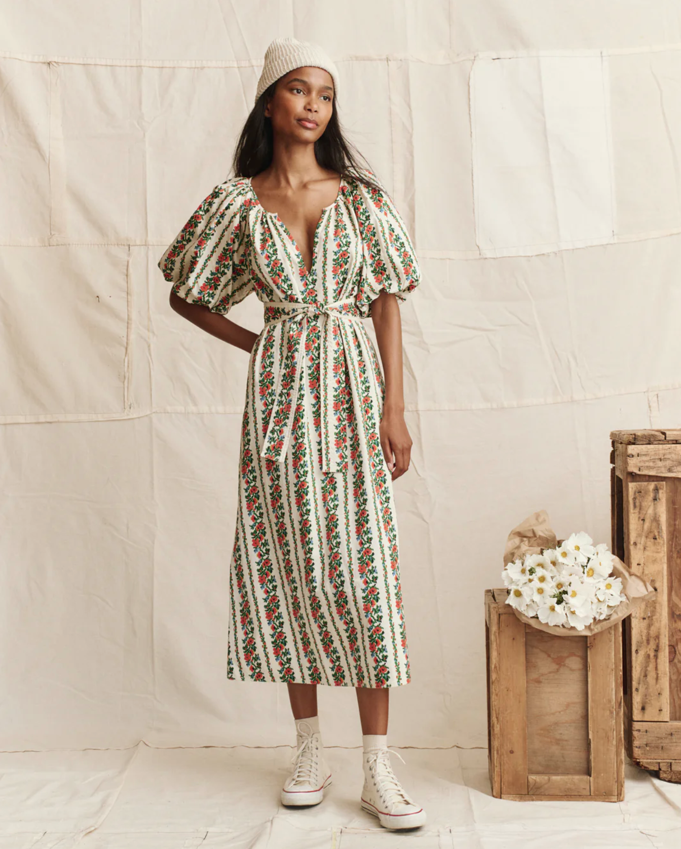 A woman in The Great Inc.'s vintage cotton Fairground Dress with floral stripe puff sleeves, a white knit hat, and high-top sneakers stands indoors by wooden crates and white flowers, against a backdrop of cream fabric.