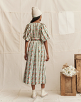 A person stands facing away, wearing The Fairground Dress by The Great Inc., featuring a western lace print and puff sleeves, paired with a white beanie and sneakers. Beside them are wooden crates with white flowers against a neutral backdrop.