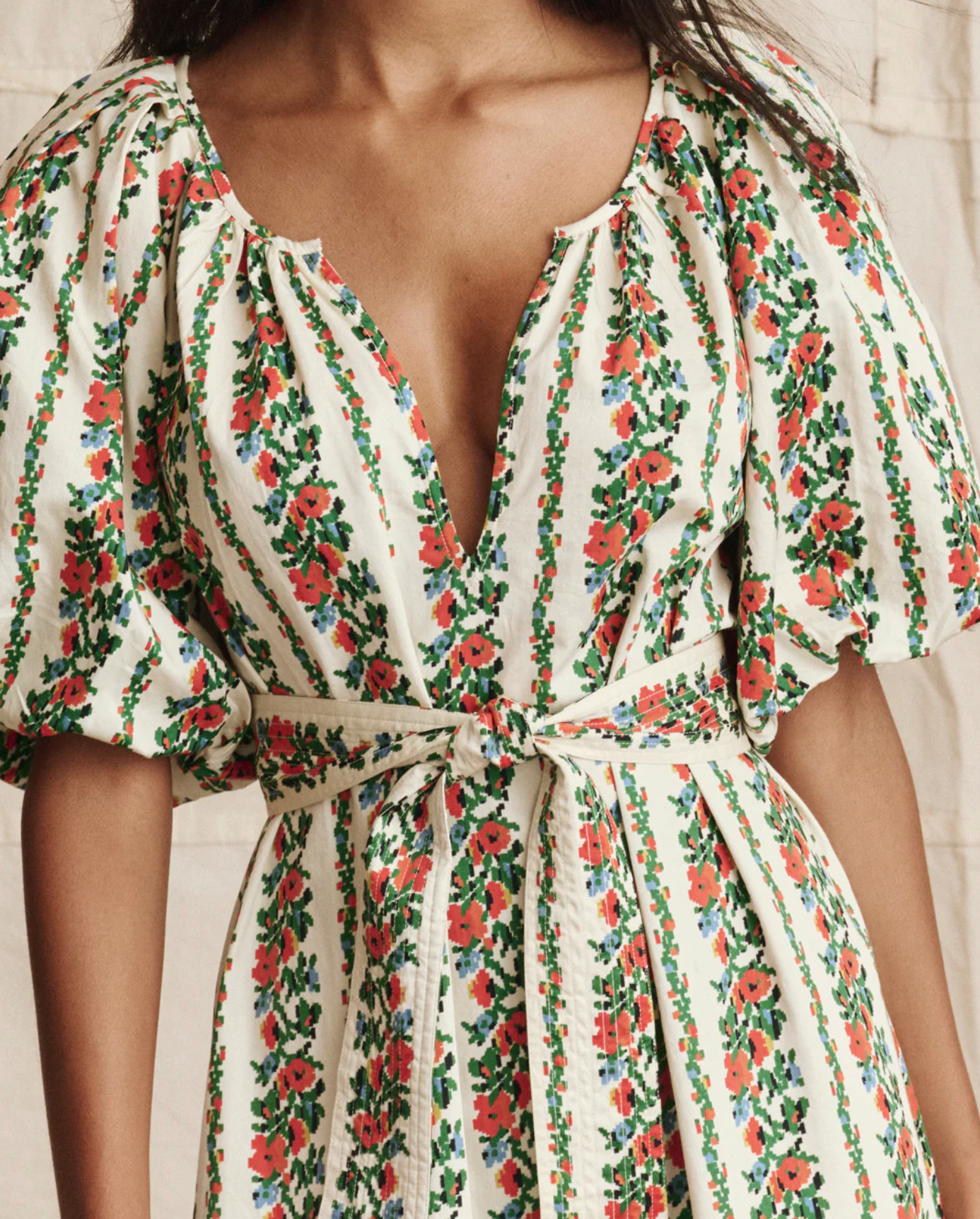 A woman wears The Fairground Dress by The Great Inc.—a green and red floral stripe pullover with puffed short sleeves, a deep V-neckline, and a matching fabric belt at the waist.