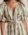 A woman wears The Fairground Dress by The Great Inc.—a green and red floral stripe pullover with puffed short sleeves, a deep V-neckline, and a matching fabric belt at the waist.