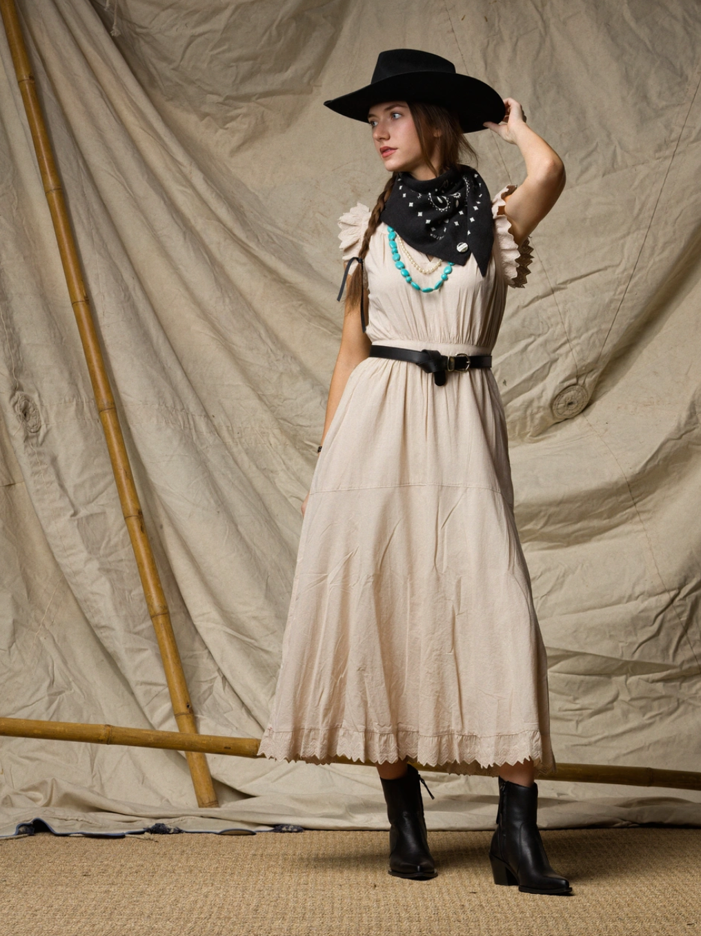 A woman stands against a beige draped backdrop, looking to the side and holding her hat. She wears the Xirena Britta Dress, styled with black boots, a wide-brimmed black hat, and a black bandana accented with turquoise beads.