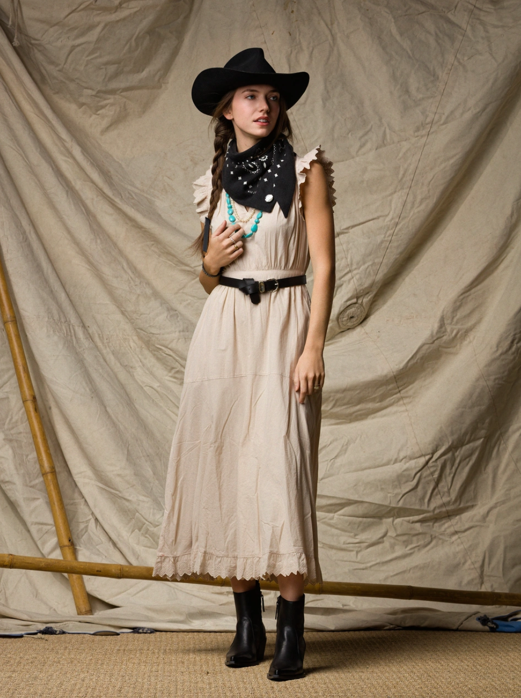A woman in the Xirena Britta Dress, styled with a black cowboy hat, boots, bandana, and turquoise necklace, stands before a beige backdrop with a bamboo pole for a Western-inspired look.