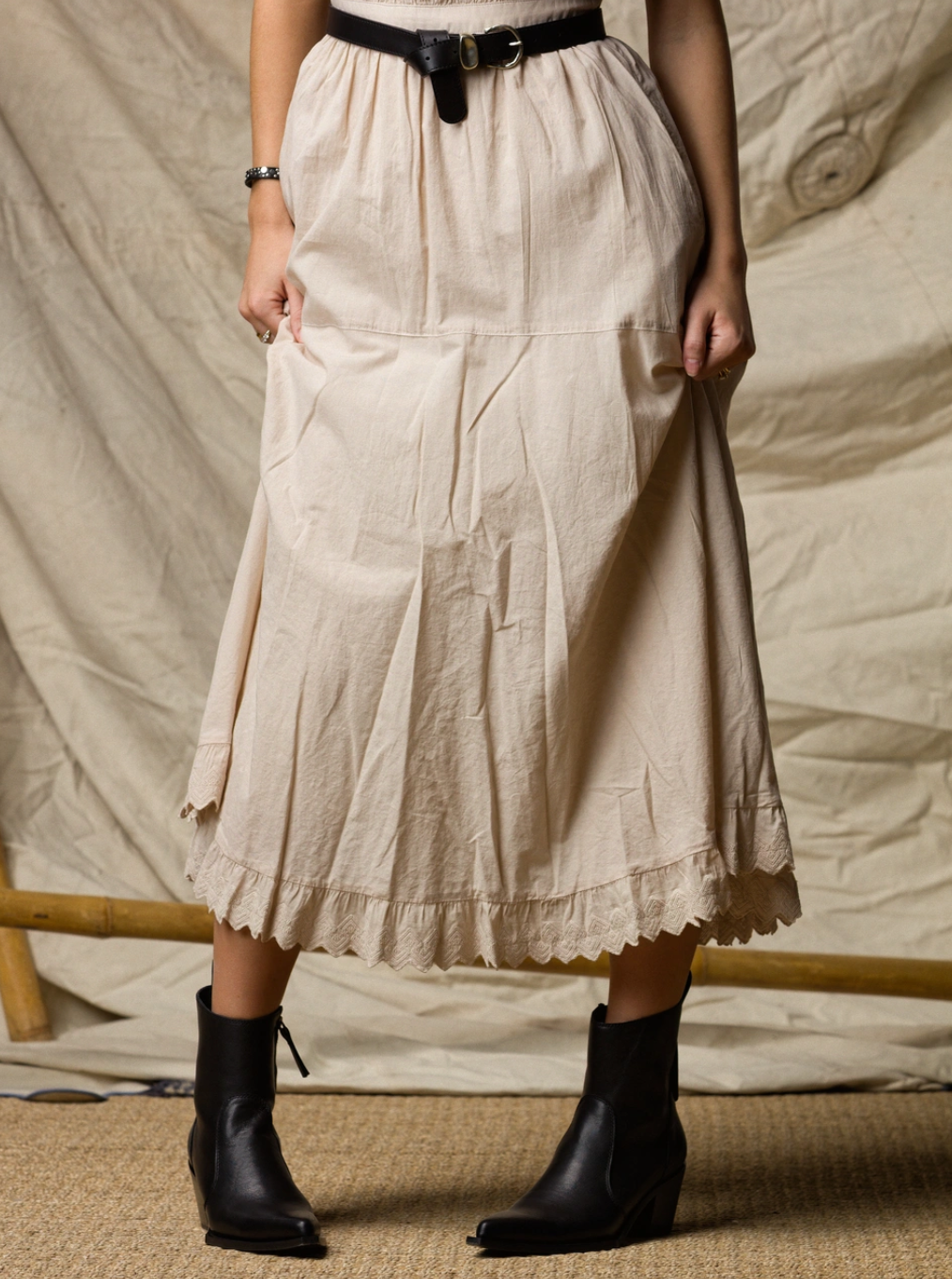 A person wearing the Xirena Britta Dress stands against a neutral, textured fabric backdrop, holding the sides of the long, light beige skirt detailed with lace and ruffles. The look is styled with a black belt and black ankle boots.