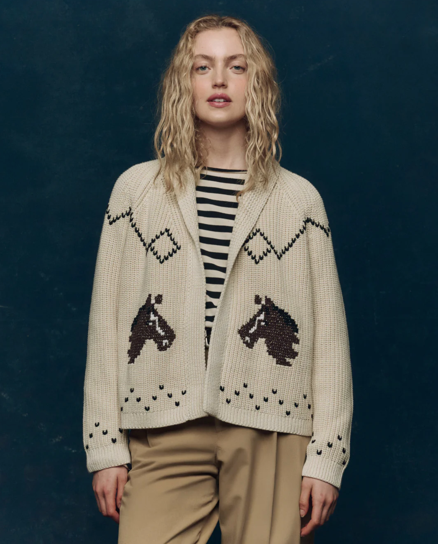 A person with wavy blonde hair wears The Horse Lodge Cardigan by The Great Inc., with hand-embroidered horses and zigzag pattern, over a black-and-white striped shirt and beige pants, standing against a dark blue background.