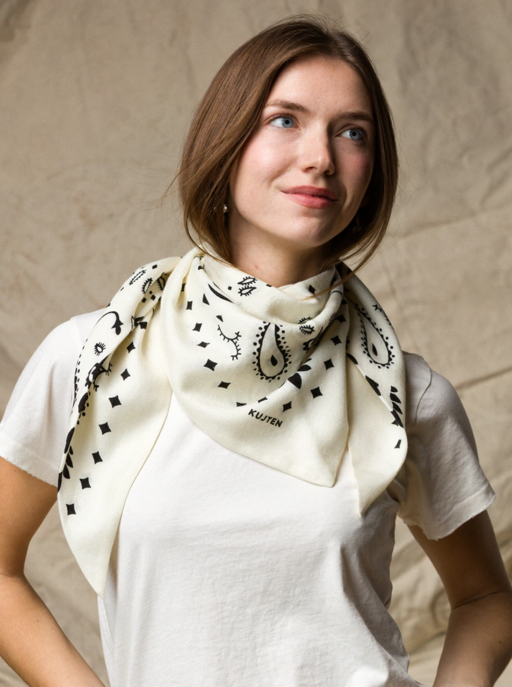 A woman wears the KUJTEN TIFA Scarf, featuring black patterns on cream, styled with a white t-shirt. She stands against a beige background, gazing softly to the side with a gentle smile.