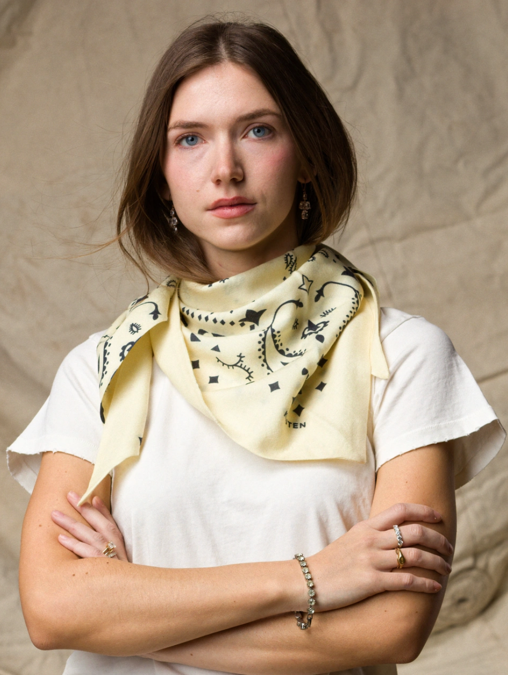 A woman with light brown hair crosses her arms and gazes forward, wearing a KUJTEN TIFA Scarf with black patterns over a white t-shirt, standing against a neutral, textured fabric backdrop.